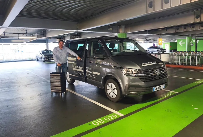 Le chauffeur Transf’Air, en tenue soignée, avec une valise devant le véhicule, sur le parking de l’aéroport.